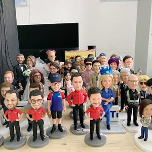 May include: A collection of bobblehead figurines, each depicting a person with unique features and attire. The figures vary in clothing, including uniforms, suits, and casual wear. They stand on individual circular bases, creating a diverse display.