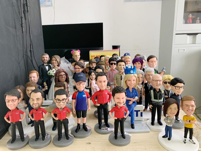 May include: A collection of bobblehead figurines, each depicting a person with unique features and attire. The figures vary in clothing, including uniforms, suits, and casual wear. They stand on individual circular bases, creating a diverse display.