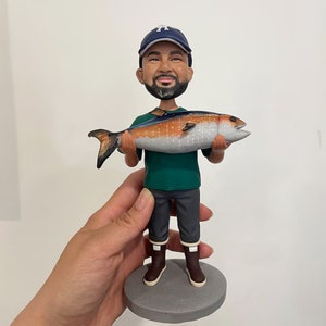 May include: A figurine of a man wearing a baseball cap and green shirt, holding a large fish. The fish is orange and silver with black fins. The man is wearing gray pants and brown boots. The figurine is on a gray base.