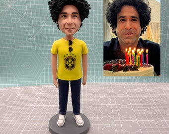 Puede incluir: Una figura personalizada de una persona con cabello castaño rizado que lleva una camiseta amarilla con el logotipo de un equipo de fútbol brasileño y pantalones vaqueros azules. La figura está de pie sobre una base gris.