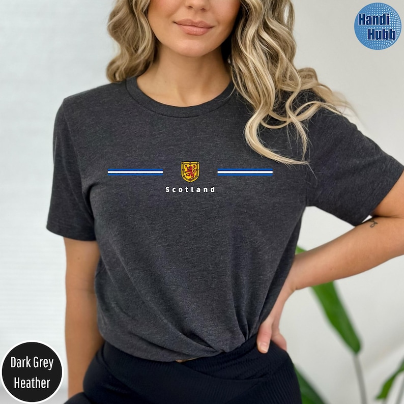 May include: Dark grey heather t-shirt with a graphic design featuring a blue stripe, a gold and red emblem, and the word "Scotland" in white. The shirt is a crew neck style.