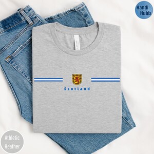 May include: Athletic heather gray t-shirt with a crew neck. The shirt features a horizontal design with blue stripes and a gold and red emblem, with the word "Scotland" below. The shirt is folded on top of blue jeans.