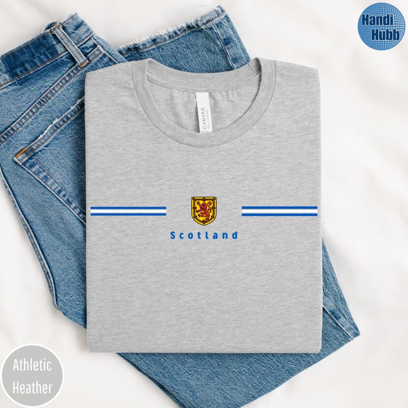 May include: Athletic heather gray t-shirt with a crew neck. The shirt features a horizontal design with blue stripes and a gold and red emblem, with the word "Scotland" below. The shirt is folded on top of blue jeans.