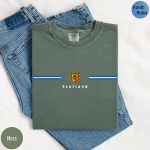May include: Moss green t-shirt with a horizontal blue and white stripe design and a Scottish emblem. The word "Scotland" is printed below the emblem. The shirt is folded on top of a pair of blue jeans.