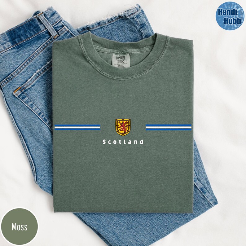 May include: Moss green t-shirt with a horizontal blue and white stripe design and a Scottish emblem. The word "Scotland" is printed below the emblem. The shirt is folded on top of a pair of blue jeans.