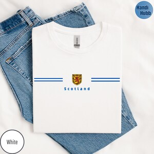 May include: White t-shirt with a crew neck, featuring a Scottish emblem design. The design includes a yellow and red shield with a lion, two blue horizontal stripes, and the word "Scotland" in blue. The shirt is folded on blue jeans.