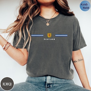 May include: Dark gray t-shirt with a crew neck featuring a design with the word "Scotland" in white, a gold and red emblem, and two blue stripes. The shirt is made of a soft, comfortable material.