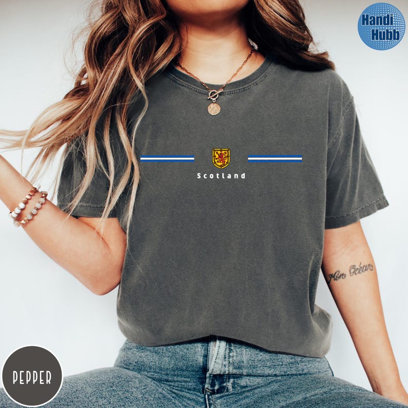 May include: Dark gray t-shirt with a crew neck featuring a design with the word "Scotland" in white, a gold and red emblem, and two blue stripes. The shirt is made of a soft, comfortable material.