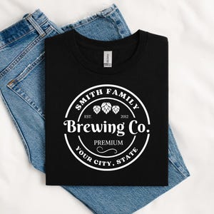 May include: A black t-shirt featuring a white circular graphic with the text Smith Family Brewing Co. Est. 2012 Premium Your City, State and three hop illustrations. The shirt is folded and placed on top of a pair of blue denim jeans.