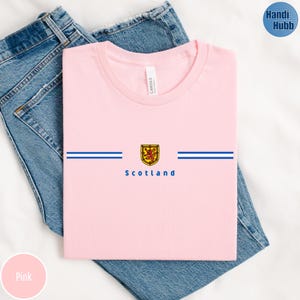 May include: Pink t-shirt with a crew neck, featuring a Scottish emblem and the word "Scotland" in blue. Two horizontal blue stripes are above and below the emblem. The shirt is folded on top of blue jeans.