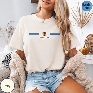 May include: Ivory-colored t-shirt with a blue and gold emblem and the word "Scotland" printed below. Two horizontal blue lines are on either side of the emblem. The shirt has short sleeves and a crew neck.
