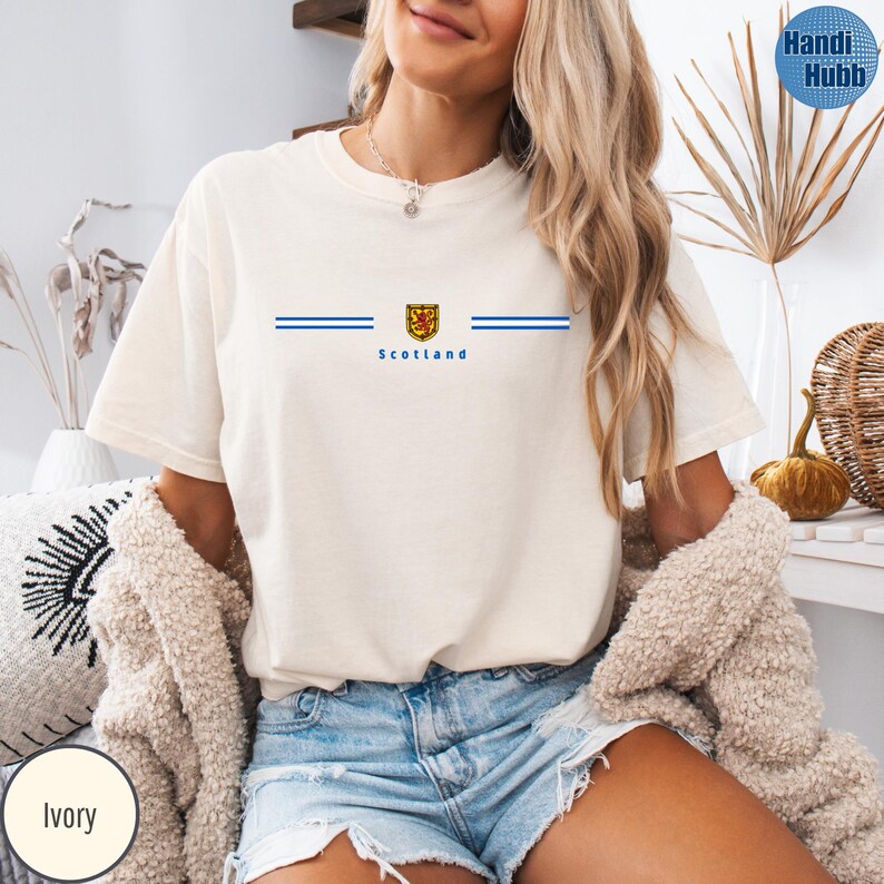 May include: Ivory-colored t-shirt with a blue and gold emblem and the word "Scotland" printed below. Two horizontal blue lines are on either side of the emblem. The shirt has short sleeves and a crew neck.