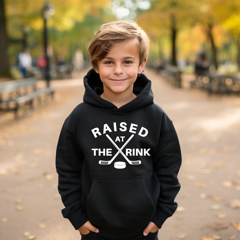 Może przedstawiać: Czarna bluza z kapturem z białym napisem "RAISED AT THE RINK". Projekt zawiera skrzyżowane kije hokejowe i krążek. Bluza ma przednią kieszeń i kaptur ze sznurkiem.