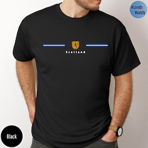 May include: Black t-shirt featuring the word "Scotland" in white, with a gold and red emblem and two horizontal blue and white stripes. The shirt has a crew neck and short sleeves.
