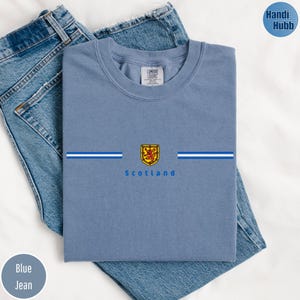 May include: A light blue t-shirt with the word "Scotland" printed below a crest. The crest is gold, red, and yellow. The shirt has two horizontal white and blue stripes. The shirt is folded on top of blue jeans.