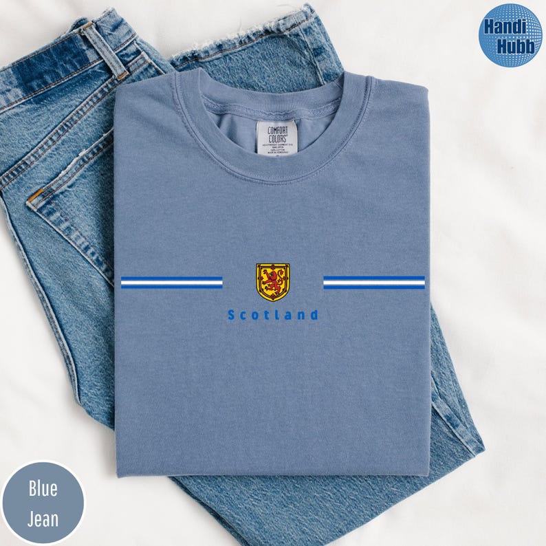 May include: A light blue t-shirt with the word "Scotland" printed below a crest. The crest is gold, red, and yellow. The shirt has two horizontal white and blue stripes. The shirt is folded on top of blue jeans.
