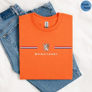 May include: Orange t-shirt with the word "Netherlands" printed in white. The shirt features a design with a lion emblem and horizontal stripes in red, white, and blue. The shirt is folded on top of blue jeans.