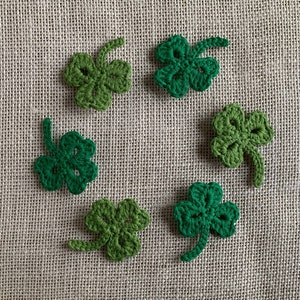 Puede incluir: Seis tréboles de cuatro hojas de ganchillo verde dispuestos en círculo sobre un fondo de arpillera beige.