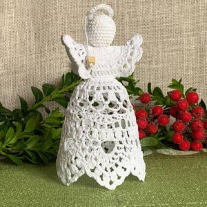 May include: White crocheted angel ornament with a bell-shaped skirt and a halo. The angel is holding a small wooden piece in its hands. The ornament is sitting on a green surface with red berries and green leaves in the background.