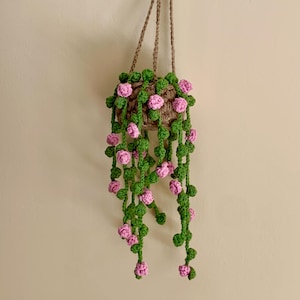 May include: A crocheted hanging planter with pink flowers and green leaves. The planter is made of brown twine and has a rustic look.