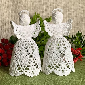 Puede incluir: Dos adornos de ángel de crochet blanco con detalles de encaje. Los ángeles están de pie sobre una superficie verde con bayas rojas.