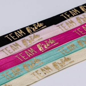 Könnte beinhalten: Nahaufnahme von mehreren elastischen Stirnbändern in verschiedenen Farben, darunter Schwarz, Rosa und Türkis. Jedes Stirnband ist mit dem goldenen Text "TEAM Bride" in einer Schreibschrift bedruckt, ideal für einen Junggesellinnenabschied.