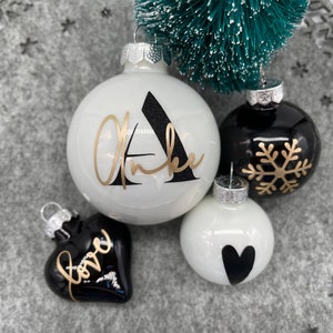 Könnte beinhalten: Eine Sammlung von Weihnachtsornamenten in Schwarz und Weiß. Ein weißer Ornament hat einen schwarzen Buchstaben "A" und goldene Schrift. Weitere Ornamente sind ein schwarzes Herz mit "love" in Gold, eine schwarze Kugel mit einem goldenen Schneeflocke und eine weiße Kugel mit einem schwarzen Herz.