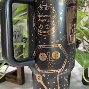 May include: Black travel mug with a gold metallic finish. The mug features a variety of gold designs including stars, a key, buttons, and the text "Welcome home!" and "Hey Jinxy, look what I found in Grandma's trunk. Look familiar? Wybie"