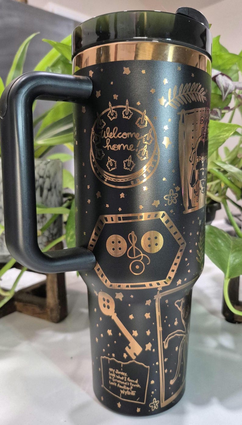 May include: Black travel mug with a gold metallic finish. The mug features a variety of gold designs including stars, a key, buttons, and the text "Welcome home!" and "Hey Jinxy, look what I found in Grandma's trunk. Look familiar? Wybie"
