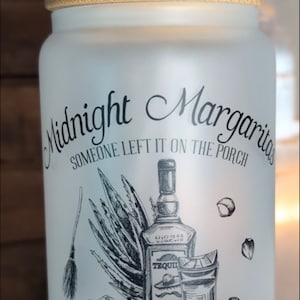 May include: White ceramic mug with a black and white illustration of a bottle of tequila, a glass of margarita, a frog, and a cactus. The mug has the text "Midnight Margaritas Someone Left It On The Porch Be Careful What You Wish For Founded Since 1998".