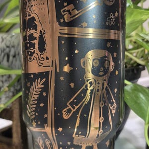 May include: Black stainless steel tumbler with a gold design featuring characters and objects from the movie Coraline. The design includes a key, a button eye, a ghost, and the text "Coraline".