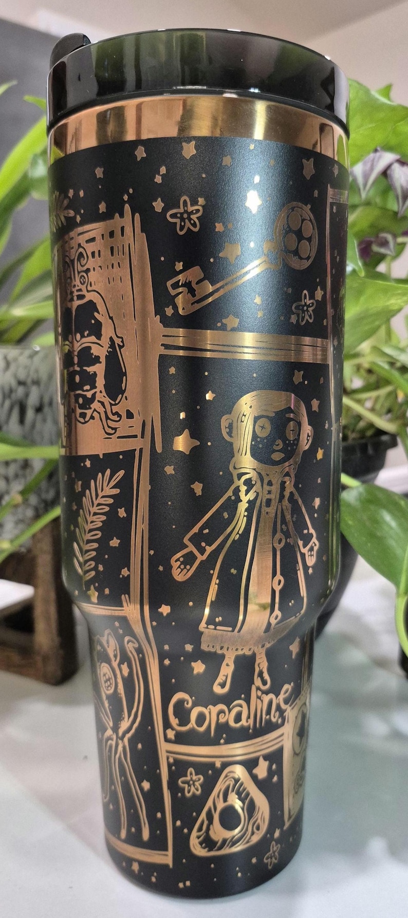 May include: Black stainless steel tumbler with a gold design featuring characters and objects from the movie Coraline. The design includes a key, a button eye, a ghost, and the text "Coraline".