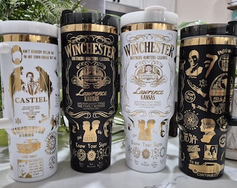May include: Four travel mugs with black and gold or white and gold designs. The mugs feature the text "Winchester" and "Lawrence Kansas", along with various illustrations and quotes. One mug has a handle and the text "Castiel".