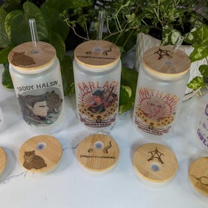 May include: Set of six frosted glass tumblers with bamboo lids and straws. Each tumbler features a different design with text and illustrations. The designs include "Boost Arion", "Daddy Halsin", "Harlaquin's Gate", "Mintha", "Jo My Girlfriend", and a cat design.