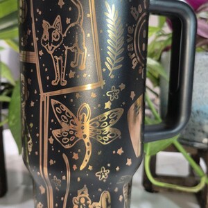 May include: Black travel mug with a gold metallic finish. The mug features a variety of gold designs including a cat, a dragonfly, a fern, and the words "Be Brave".