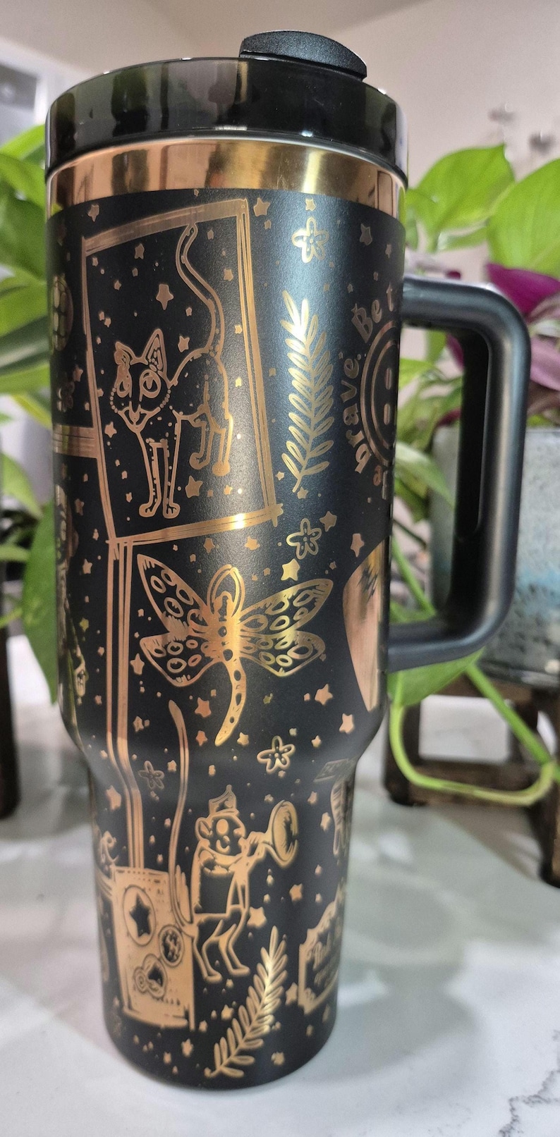 May include: Black travel mug with a gold metallic finish. The mug features a variety of gold designs including a cat, a dragonfly, a fern, and the words "Be Brave".