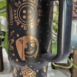 May include: Black stainless steel travel mug with a gold-colored design featuring a button, a character with lightning bolts, and the text "Be brave. Be tricky. Be wise." The mug has a black handle and a gold-colored logo that says "Pink Palace Apartments."