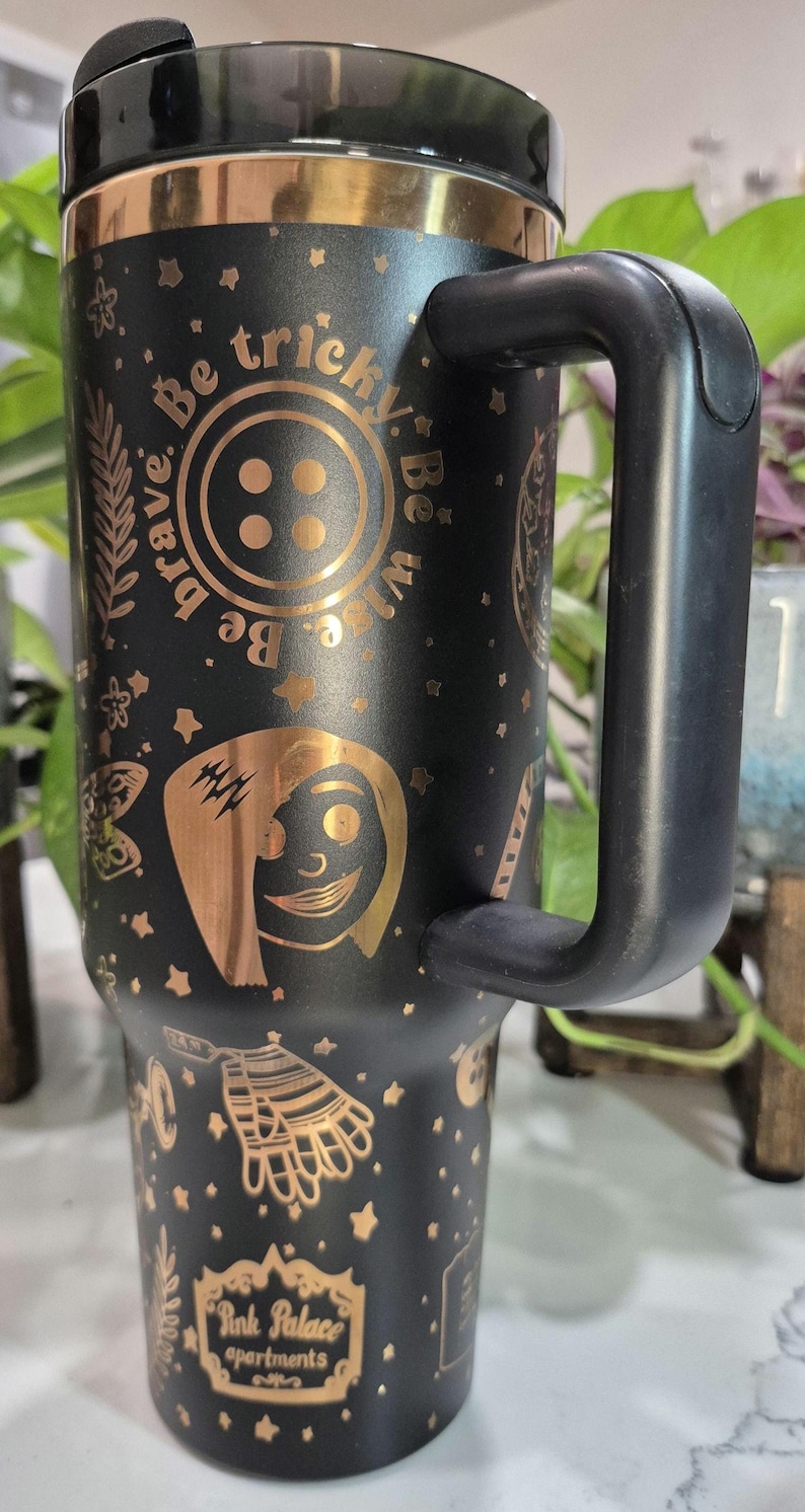 May include: Black stainless steel travel mug with a gold-colored design featuring a button, a character with lightning bolts, and the text "Be brave. Be tricky. Be wise." The mug has a black handle and a gold-colored logo that says "Pink Palace Apartments."