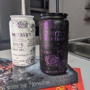 Puede incluir: Dos vasos de acero inoxidable con tapas y pajitas negras y blancas. El vaso blanco tiene un diseño en blanco y negro con el texto "MUTSLUT" y "YOU HAD ME AT TRIGGER WARNING". El vaso negro tiene un diseño en blanco y negro con el texto "DARK BOOK CLUB" y "BOYFRIEND NATIVE YOU CRAWL TO ME GOOD LITTLE SMUT".