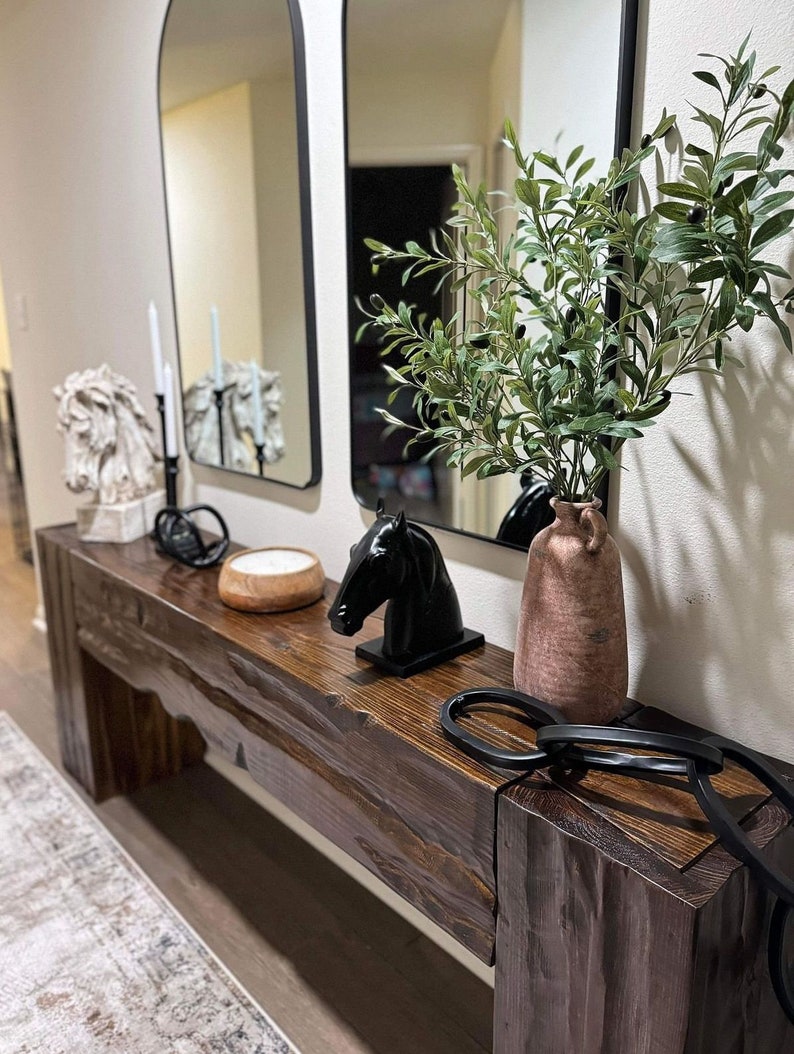 May include: A dark wood console table with a black horse head sculpture, a white candle holder, a white ceramic horse head sculpture, a vase with green leafy branches, and a black ring on the surface.