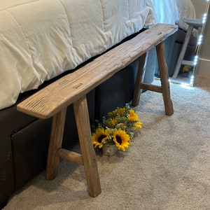 May include: A rustic wooden bench with a natural finish sits next to a bed with a white comforter. The bench has a simple design with four legs and a flat top. A small potted plant with yellow flowers sits in front of the bench.