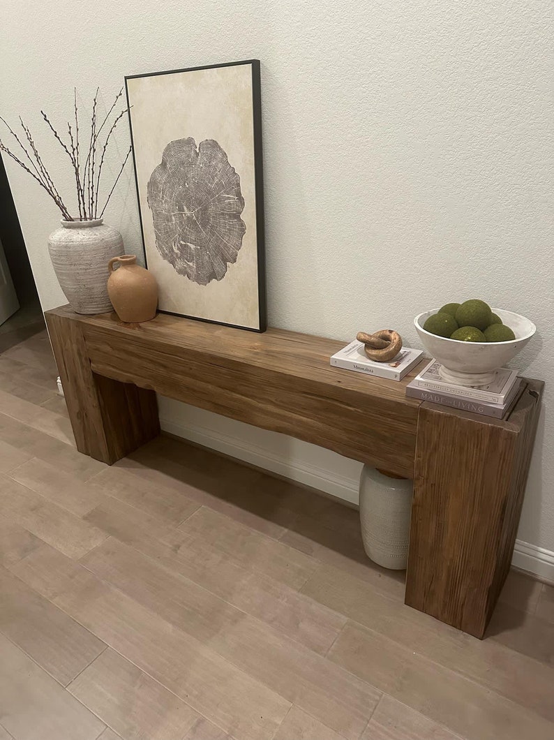 May include: A rustic wooden console table with a thick, natural wood top and two sturdy legs. The table is decorated with a vase of dried branches, a ceramic vase, a small terracotta vase, a framed print of a tree ring, a bowl of green decorative balls, and a stack of books.
