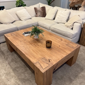 May include: A large, rectangular wooden coffee table with a natural wood finish. The table has a thick, chunky top and legs with a unique, geometric design. The table is sitting on a light brown rug.