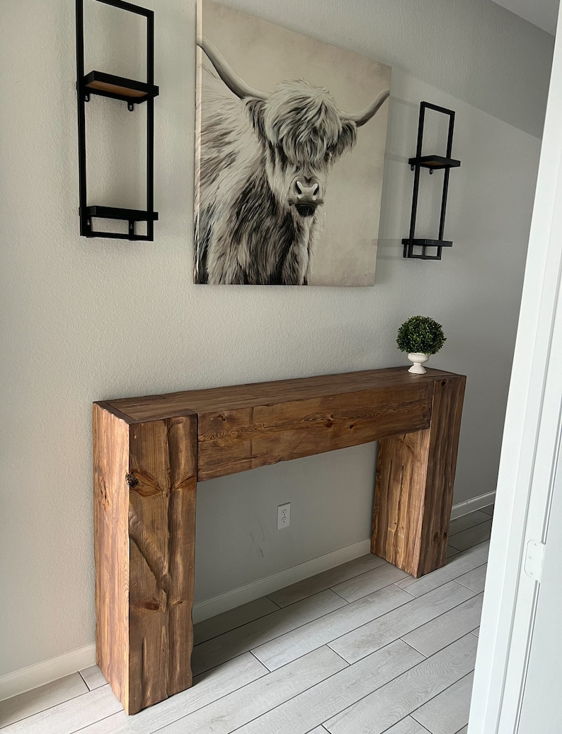 May include: A rustic wooden console table with a natural wood finish. The table has a single drawer and is supported by two thick, rectangular legs. The table is in a hallway with a white wall and a framed print of a Highland cow.