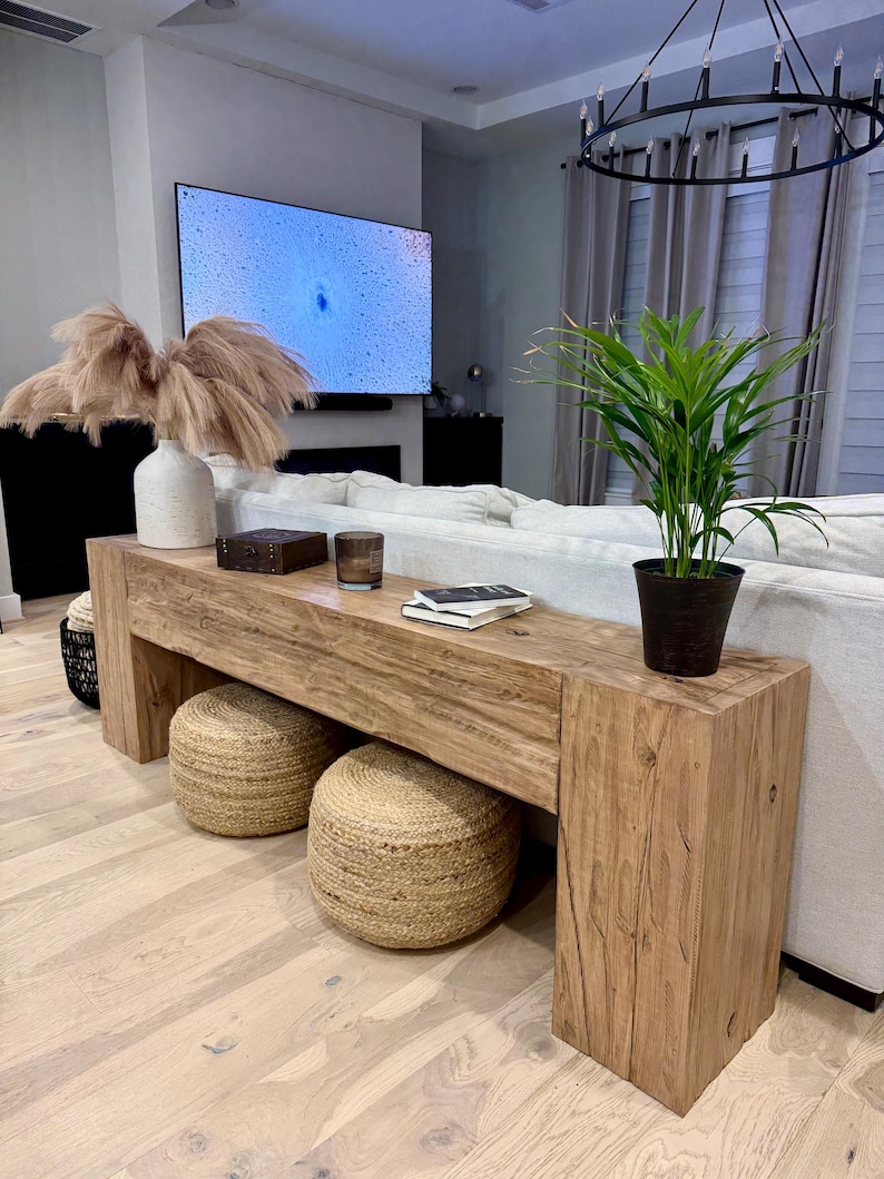 May include: A rustic wooden console table with a natural wood finish. The table has two open shelves and is decorated with a vase of dried pampas grass, a small wooden box, and two books. Two woven rope ottomans are placed in front of the table.