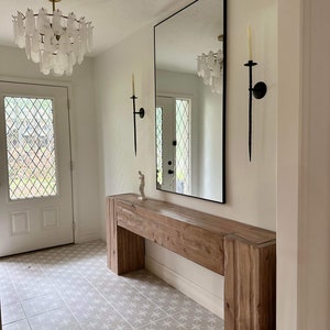 May include: A wooden console table with a natural finish and a white tiled floor. The table has a long, rectangular top and two vertical supports. A large mirror hangs on the wall behind the table, reflecting the chandelier above.