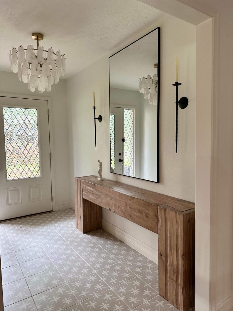 May include: A wooden console table with a natural finish and a white tiled floor. The table has a long, rectangular top and two vertical supports. A large mirror hangs on the wall behind the table, reflecting the chandelier above.
