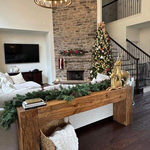 May include: A rustic wooden console table with a live edge top and a white faux fur throw in a woven basket. The table is decorated with a garland of evergreen branches and a stack of books. The table is in front of a fireplace with a stone surround and a Christmas tree decorated with ornaments and lights.
