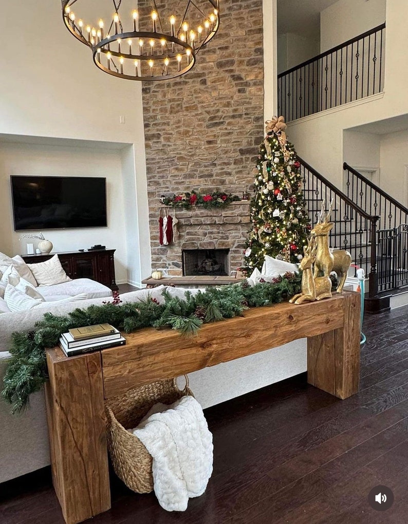May include: A rustic wooden console table with a live edge top and a white faux fur throw in a woven basket. The table is decorated with a garland of evergreen branches and a stack of books. The table is in front of a fireplace with a stone surround and a Christmas tree decorated with ornaments and lights.