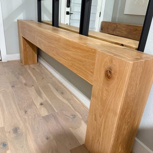 May include: A long, wooden console table with a natural wood finish. The table has a simple, rectangular design and is supported by two square legs. The table is positioned in a hallway with a mirror wall behind it.